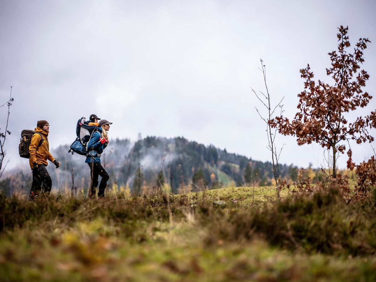 Two people hiking in a natural outdoor setting are wearing gloves and outdoor clothing. One of them is carrying a child in a backpack carrier.