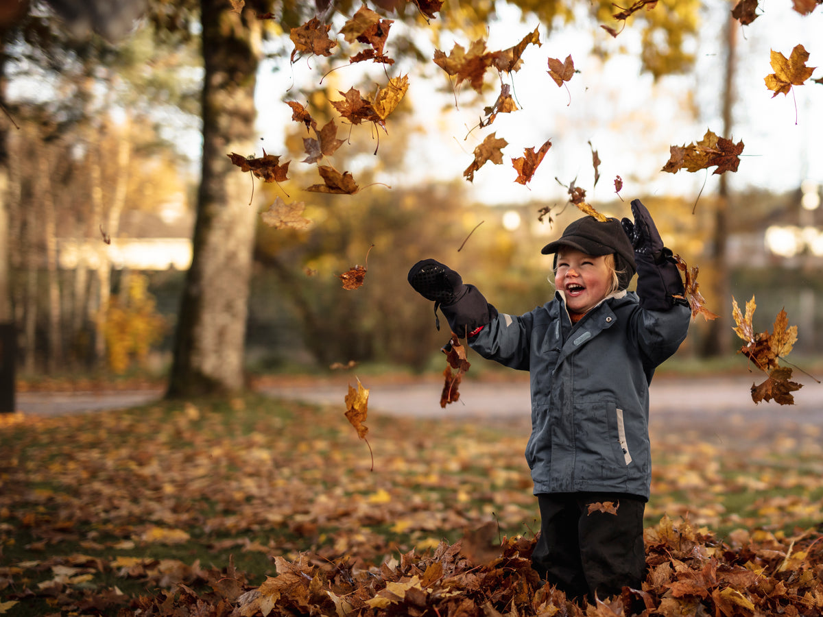 Child wearing insulated mittens and outdoor clothing, playing in falling autumn leaves outdoors.